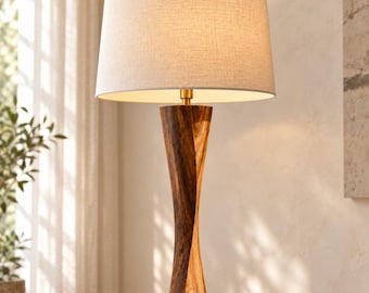 Może przedstawiać: Lampa stołowa z wysoką, spiralną podstawą z ciemnego drewna o naturalnym usłojeniu oraz cylindrycznym abażurem z jasnobeżowej tkaniny. Lampa posiada mały łącznik z metalu w kolorze złota pomiędzy drewnianą podstawą a abażurem.
