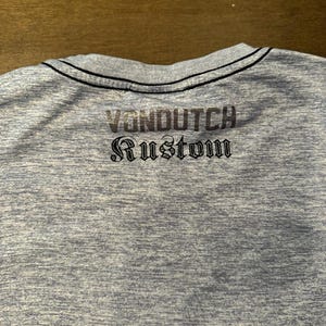 May include: A light blue t-shirt with the text "VONDUTCH Rustom" printed on the back in a dark grey colour. The shirt has a black trim around the neckline.