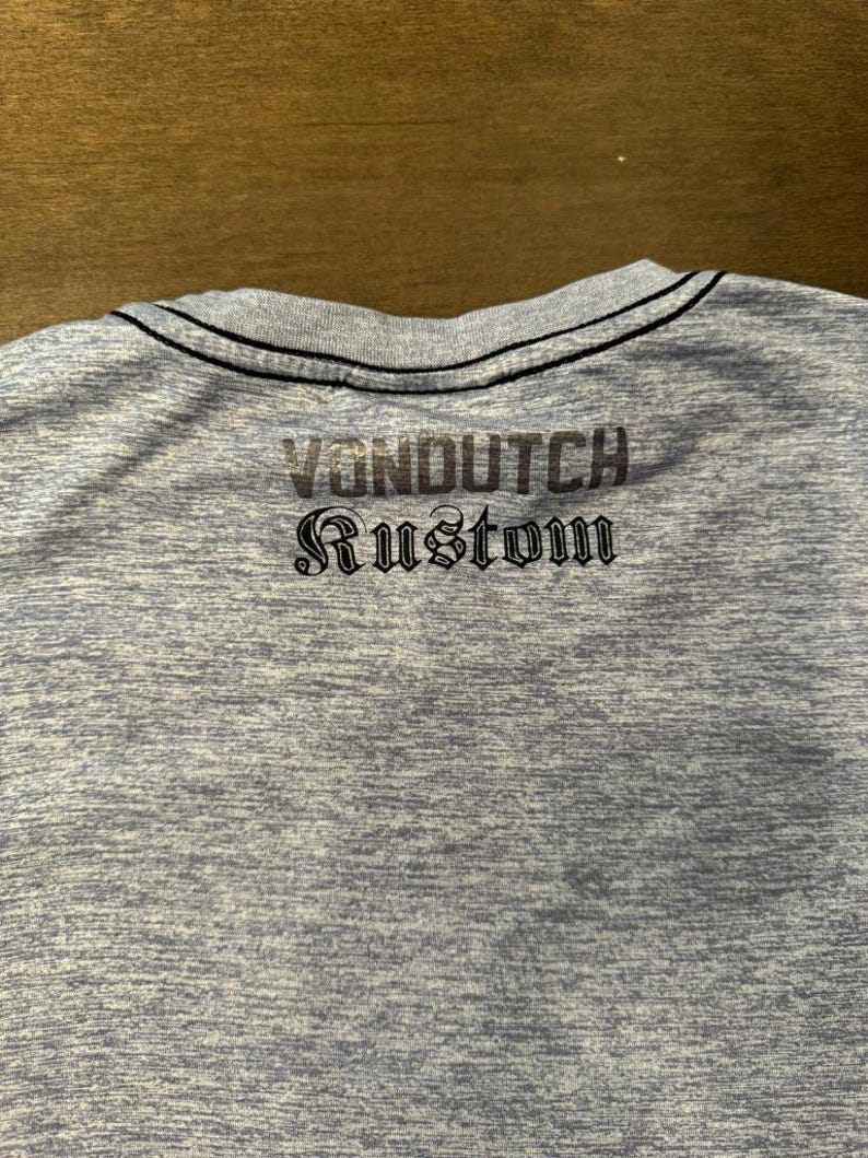 May include: A light blue t-shirt with the text "VONDUTCH Rustom" printed on the back in a dark grey colour. The shirt has a black trim around the neckline.