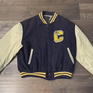 May include: Navy blue and tan varsity jacket with a yellow "C" patch that says "California". The jacket has tan sleeves, a navy blue body, and yellow and navy blue striped trim on the cuffs, collar, and hem.