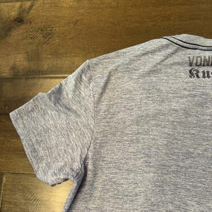 May include: A light blue short-sleeved t-shirt with the text "VON RUG" printed on the back. The shirt has a crew neck and a heathered texture.