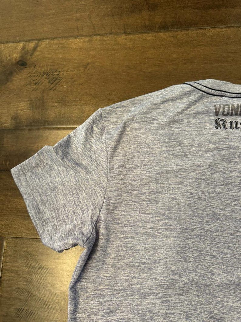 May include: A light blue short-sleeved t-shirt with the text "VON RUG" printed on the back. The shirt has a crew neck and a heathered texture.