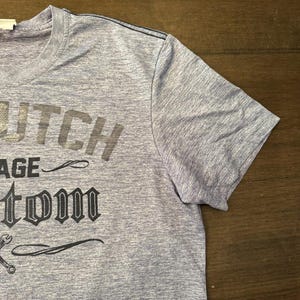 May include: A light blue t-shirt featuring the text "Dutch Garage Custom" in dark grey. The shirt has a crew neck and short sleeves. A wrench graphic is visible below the text.