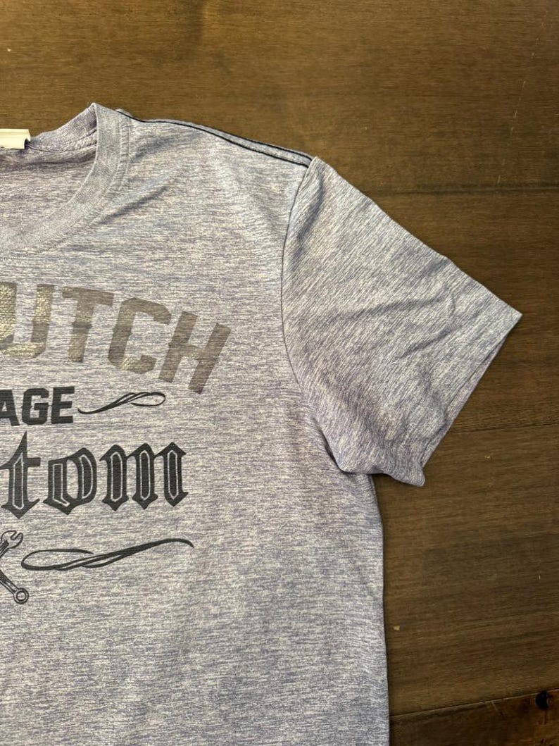 May include: A light blue t-shirt featuring the text "Dutch Garage Custom" in dark grey. The shirt has a crew neck and short sleeves. A wrench graphic is visible below the text.