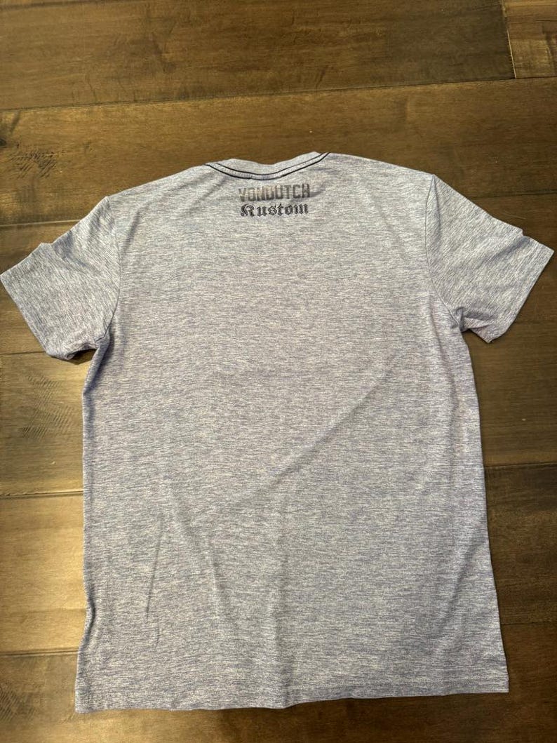 May include: A grey marl t-shirt with short sleeves and a dark blue trim around the crew neck. The text "Von Dutch Kustom" is printed across the upper back of the shirt in dark blue.