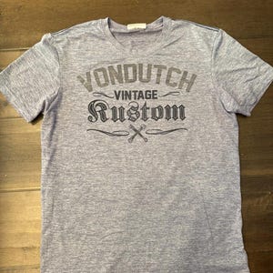 May include: A heather grey short-sleeved t-shirt featuring the text "VONDUTCH VINTAGE RUSTOM" in a distressed font. The design includes crossed wrenches. The shirt has a crew neck.
