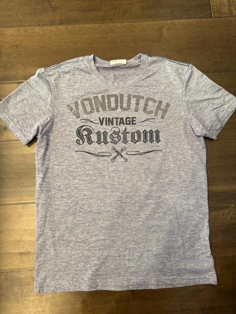 May include: A heather grey short-sleeved t-shirt featuring the text "VONDUTCH VINTAGE RUSTOM" in a distressed font. The design includes crossed wrenches. The shirt has a crew neck.