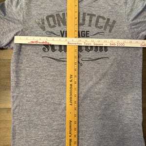 May include: A light blue t-shirt with the text "VON UTCH VIAGE" in dark grey lettering. A wooden ruler is placed vertically down the centre of the shirt, measuring approximately 74 cm. A white ruler is placed horizontally across the shirt.
