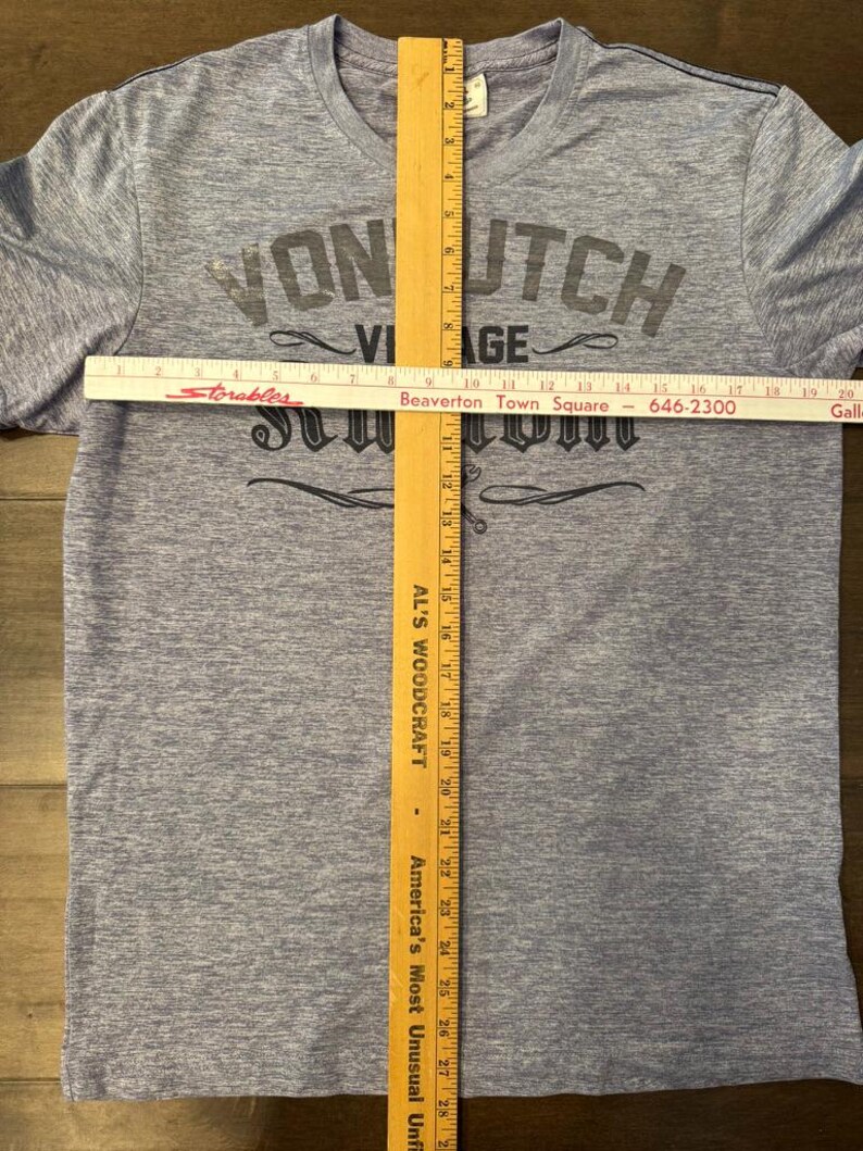May include: A light blue t-shirt with the text "VON UTCH VIAGE" in dark grey lettering. A wooden ruler is placed vertically down the centre of the shirt, measuring approximately 74 cm. A white ruler is placed horizontally across the shirt.
