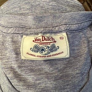 May include: Close-up of a light purple t-shirt with a ribbed neckline. The shirt features a Von Dutch label with a logo of an eye with wings and the text "FLAMES, STRIPES AND EYEBALLS". The label also has the size "XL".
