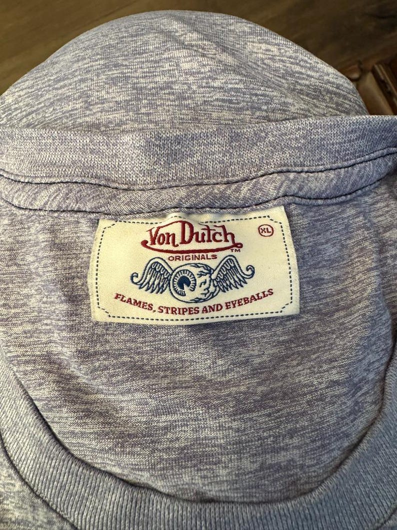 May include: Close-up of a light purple t-shirt with a ribbed neckline. The shirt features a Von Dutch label with a logo of an eye with wings and the text "FLAMES, STRIPES AND EYEBALLS". The label also has the size "XL".