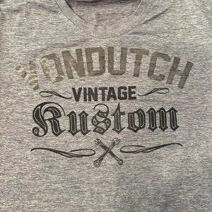 May include: A light blue V-neck t-shirt featuring the text "VON DUTCH VINTAGE RUSTOM" in dark grey. The design includes crossed wrenches, hinting at a vintage or automotive style.