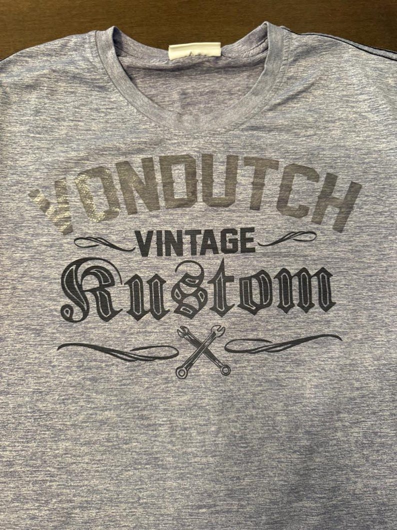 May include: A light blue V-neck t-shirt featuring the text "VON DUTCH VINTAGE RUSTOM" in dark grey. The design includes crossed wrenches, hinting at a vintage or automotive style.
