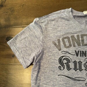 May include: A blue marl short-sleeved t-shirt featuring the words "VOND" and "RUST" in black. The shirt has a crew neck and a classic fit. The text is in a vintage style.