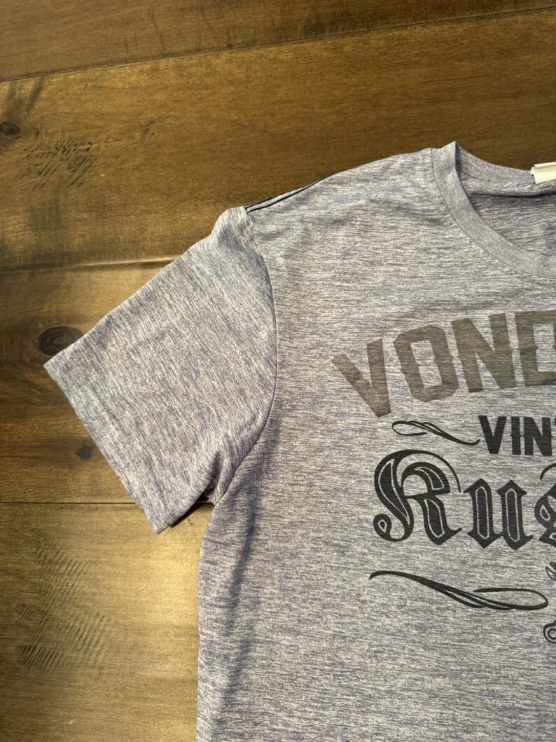 May include: A blue marl short-sleeved t-shirt featuring the words "VOND" and "RUST" in black. The shirt has a crew neck and a classic fit. The text is in a vintage style.