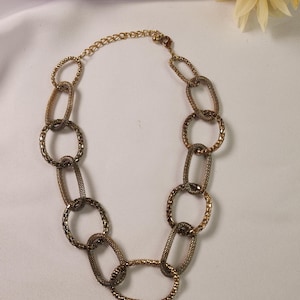 May include: A gold-toned chain necklace featuring large, interlocking oval links with a textured, mesh-like surface. The necklace includes a thin adjustable chain segment with a small lobster claw clasp closure at the top.