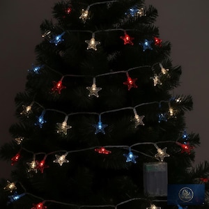 May include: A string of star-shaped LED lights featuring red, white, and blue bulbs draped across a dark green artificial Christmas tree. The lights are connected by a thin wire and include a rectangular battery pack at the bottom right.