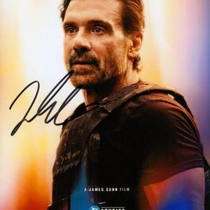 May include: A promotional poster featuring a man with dark hair and a beard wearing a black tactical vest. The text reads A JAMES GUNN FILM, DC STUDIOS, and SUPERMAN. A black ink signature is overlaid on the left side of the image.
