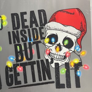 Pu&ograve; includere: Una maglietta grigia con un teschio nero che indossa un cappello da Babbo Natale rosso e circondato da luci di Natale colorate. Il testo "Dead Inside But Gettin' Lit" &egrave; stampato sulla maglietta.