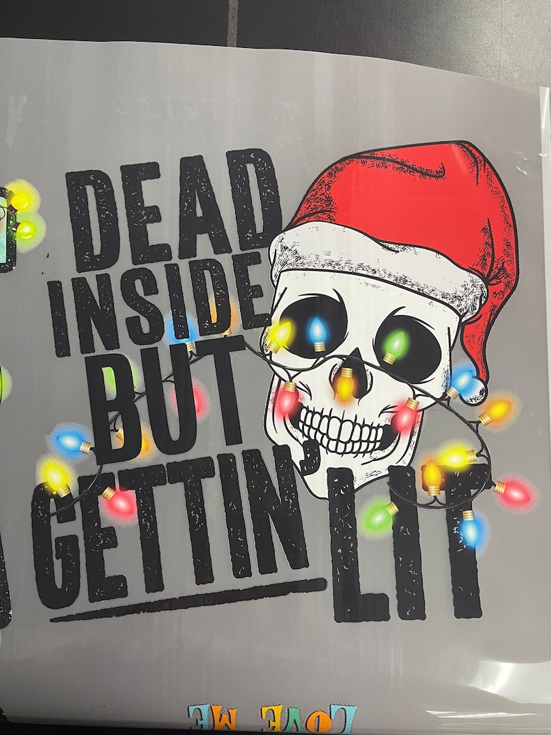 Pu&ograve; includere: Una maglietta grigia con un teschio nero che indossa un cappello da Babbo Natale rosso e circondato da luci di Natale colorate. Il testo "Dead Inside But Gettin' Lit" &egrave; stampato sulla maglietta.