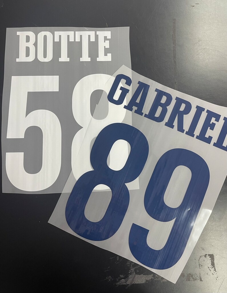 Pu&ograve; includere: Due trasferimenti a caldo bianchi con il testo "BOTTE" e i numeri "58" su uno e il testo "GABRIEL" e i numeri "89" sull'altro.