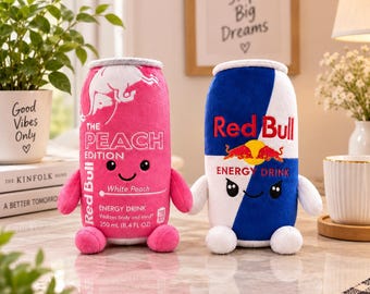 Peut inclure: Deux peluches en forme de canettes de boisson énergisante. Celle de gauche est rose avec le texte blanc The Peach Edition Red Bull Energy Drink 250 ml. Celle de droite est bleue et blanche avec le texte rouge Red Bull Energy Drink.