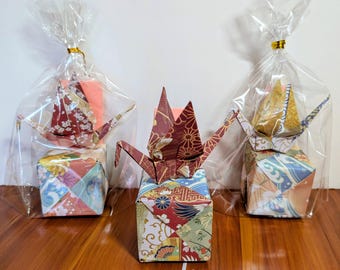May include: Three origami cranes perched on patterned paper cubes. Two cranes are enclosed in clear plastic bags with gold ties, while the center crane is displayed openly. The paper features intricate floral and wave motifs.