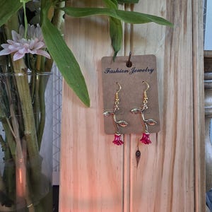 Könnte beinhalten: Ein Paar goldfarbene Hängeohrringe mit einem Rosenstiel-Design und einer roten Blütenknospe am unteren Ende. Die Ohrringe sind an einer braunen Pappkarte befestigt, auf der der Text Fashion Jewelry in Schwarz gedruckt ist.