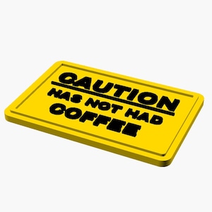 May include: A rectangular yellow sign with a raised border and black embossed text that reads CAUTION HAS NOT HAD COFFEE. The sign is oriented horizontally and features a bold, block-style font for the lettering against the bright yellow background.