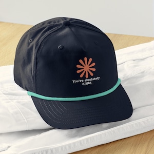May include: A navy blue baseball cap featuring a five-panel construction and a teal rope detail across the base of the crown. The front displays a terracotta-colored flower graphic above the white text that reads, You're absolutely right.
