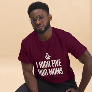 May include: A maroon short-sleeved t-shirt featuring a white graphic of a paw print with a small heart above it. Below the graphic, the text reads "I HIGH FIVE DOG MUMS" in a bold, sans-serif font. The shirt has a crew neckline.