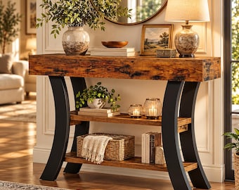 May include: A console table with a rectangular wood-grain top and two lower shelves supported by curved black metal legs. The table holds a ceramic vase with greenery, a lamp, books, a woven basket with a cloth, and glass candle holders.