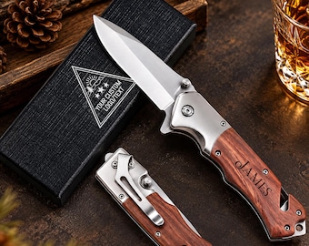 Può includere: Un coltello pieghevole con lama argentata e manico in legno venato inciso con il nome JAMES. Un secondo coltello chiuso con clip da tasca in metallo si trova sotto, accanto a una scatola regalo rettangolare nera con un logo triangolare bianco.