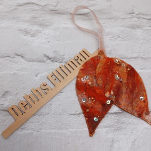 May include: A decorative felt leaf ornament in autumnal orange and brown hues, adorned with sequins and beads. It has a ribbon for hanging. A wooden sign with the text "Debbs Flliman" is also present.