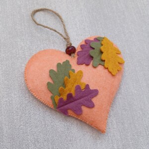 May include: A felt heart-shaped ornament with a brown twine hanger and a red bead. The heart is decorated with felt leaves in shades of green, yellow, and purple.