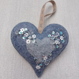 May include: A blue felt heart-shaped ornament with a white felt heart in the centre. The ornament is decorated with silver and blue sequins and white flowers.