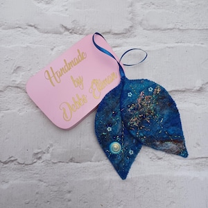 May include: A handcrafted blue felt leaf decoration, adorned with beads and sequins. A pink tag, inscribed with "Handmade by Debbie Elliman", is attached with a blue ribbon. The item is displayed against a white brick wall.