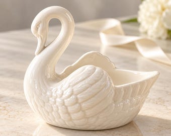 Puede incluir: Un cuenco de cerámica color crema con forma de cisne, que presenta un cuello largo y curvado y texturas de plumas moldeadas detalladas en las alas. El cisne tiene un acabado liso y brillante y un centro abierto y hueco.
