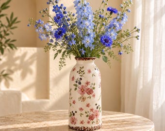 Puede incluir: Un jarrón cilíndrico de cerámica con base color crema que presenta un delicado patrón de flores y bayas en tonos rosa y verde, lleno de un vibrante arreglo de tallos altos de espuela de caballero azul y acianos.