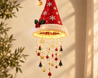 Puede incluir: Una decoración navideña colgante que presenta un gorro de Papá Noel rojo con patrones de copos de nieve blancos y un borde blanco esponjoso. Debajo, varias tiras de luces LED cálidas están adornadas con pequeños adornos rojos, verdes, estrellas y copos.