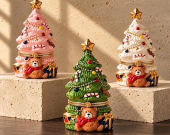 Puede incluir: Tres cajas de baratijas con bisagras en forma de árboles de Navidad en rosa, verde y blanco. Cada árbol está coronado con una estrella dorada y presenta un oso de peluche marrón con bufanda roja en la base, junto a un regalo azul con lazo dorado.