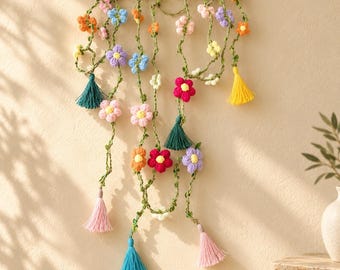 Puede incluir: Un tapiz de pared que presenta una rama de madera marrón oscuro con varias enredaderas de ganchillo verde colgando. Las enredaderas están decoradas con pequeñas flores de ganchillo de colores en tonos rosa, amarillo, naranja, azul y morado, terminadas con borlas.