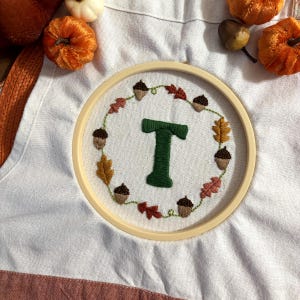 Puede incluir: Un bastidor de bordado con tela blanca y una letra T verde centrada. La letra está rodeada por una corona circular de bellotas marrones bordadas y hojas de otoño multicolores en tonos naranja, amarillo y rojo.