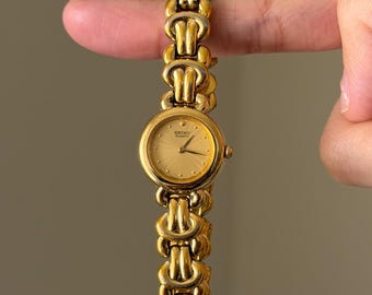 Könnte beinhalten: Eine goldfarbene Seiko Quartz Armbanduhr mit einem runden Zifferblatt mit texturiertem Strahlenmuster und einem passenden Metallgliederarmband. Das Zifferblatt zeigt den Markennamen Seiko und das Wort Quartz in schwarzer Schrift.