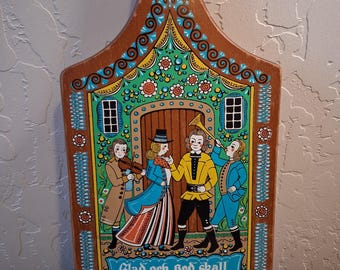 May include: A wooden cutting board with a handle and a hole, featuring a colorful painted scene of four people in historical attire. The illustration includes a violinist, a trumpet player, and text that reads: Glad och God skall människan vara.
