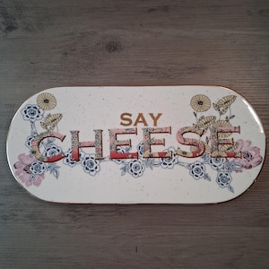 May include: An oval-shaped serving tray with a cream-colored speckled surface. The text "SAY CHEESE" is printed in the center in gold and floral-patterned letters. The tray is decorated with blue and yellow floral illustrations.