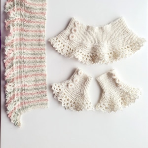 May include: A set of three knitted items in white with a delicate lace trim. The set includes a collar, leg warmers, and a scarf with pink and green stripes.