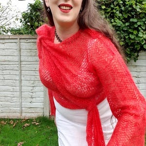 May include: A woman wearing a vibrant red, open-weave knit wrap with long sleeves. The wrap is draped over a white top and tied at the waist. The background features a wooden fence and green foliage.