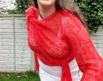 May include: A woman wearing a bright red, open-weave knit wrap with long sleeves. The wrap is draped over a white top and tied at the waist. The background includes a wooden fence and green foliage.