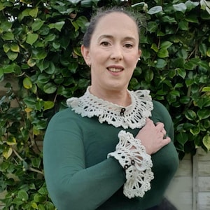 May include: A woman wearing a green long-sleeved shirt with a white crocheted collar and matching crocheted cuffs. The collar and cuffs have a scalloped edge and are decorated with small brown buttons.